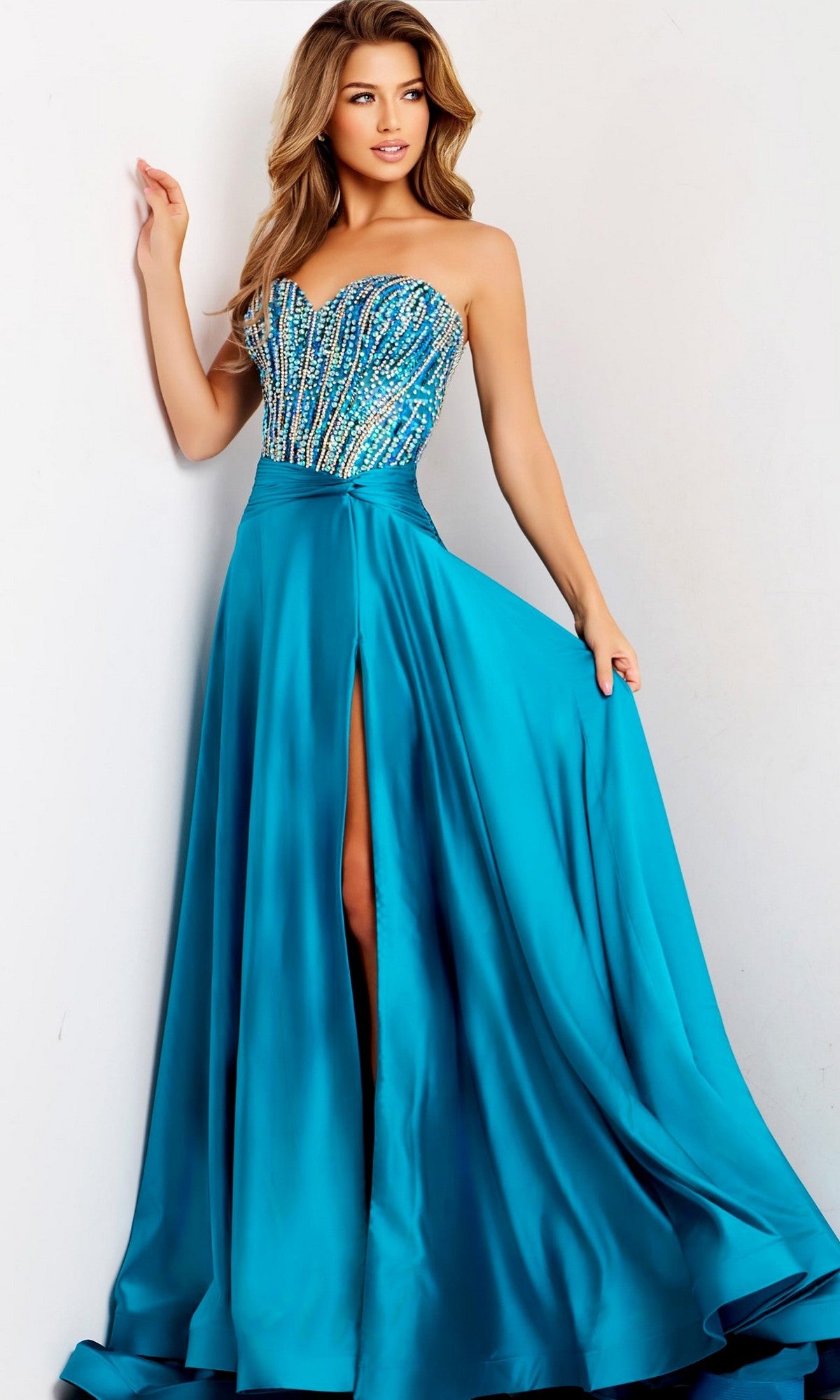 A woman wears the Jovani 43729 long prom dress in peacock, with a beaded corset bodice and an A-line satin skirt featuring a high slit, standing against a plain light-colored background.